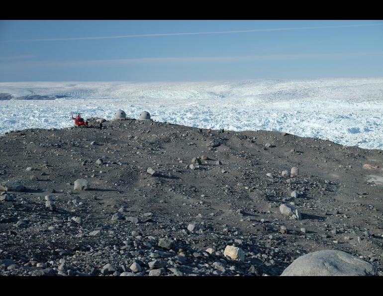 A landscape of sand and rock contrasts with the ice of Jakobshavn Glacier, one of Greenland’s fastest-moving glaciers, in 2019. A Greenland Air helicopter and research instrument installation are dwarfed by the landscape. Photo by Twila Moon/National Snow and Ice Data Center